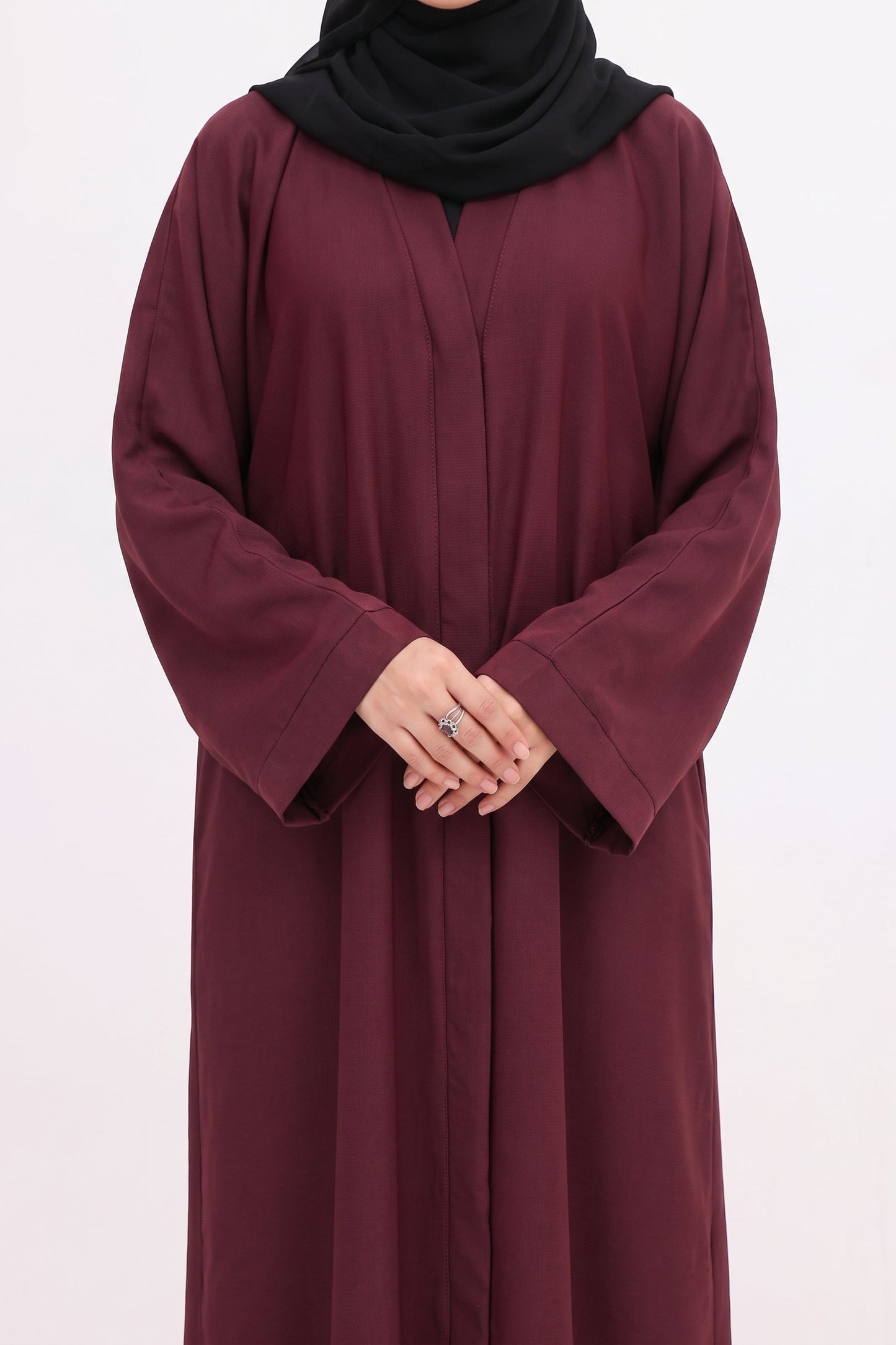 A woman wearing a long-sleeved, floor-length burgundy abaya with a black hijab, standing against a plain white studio background.