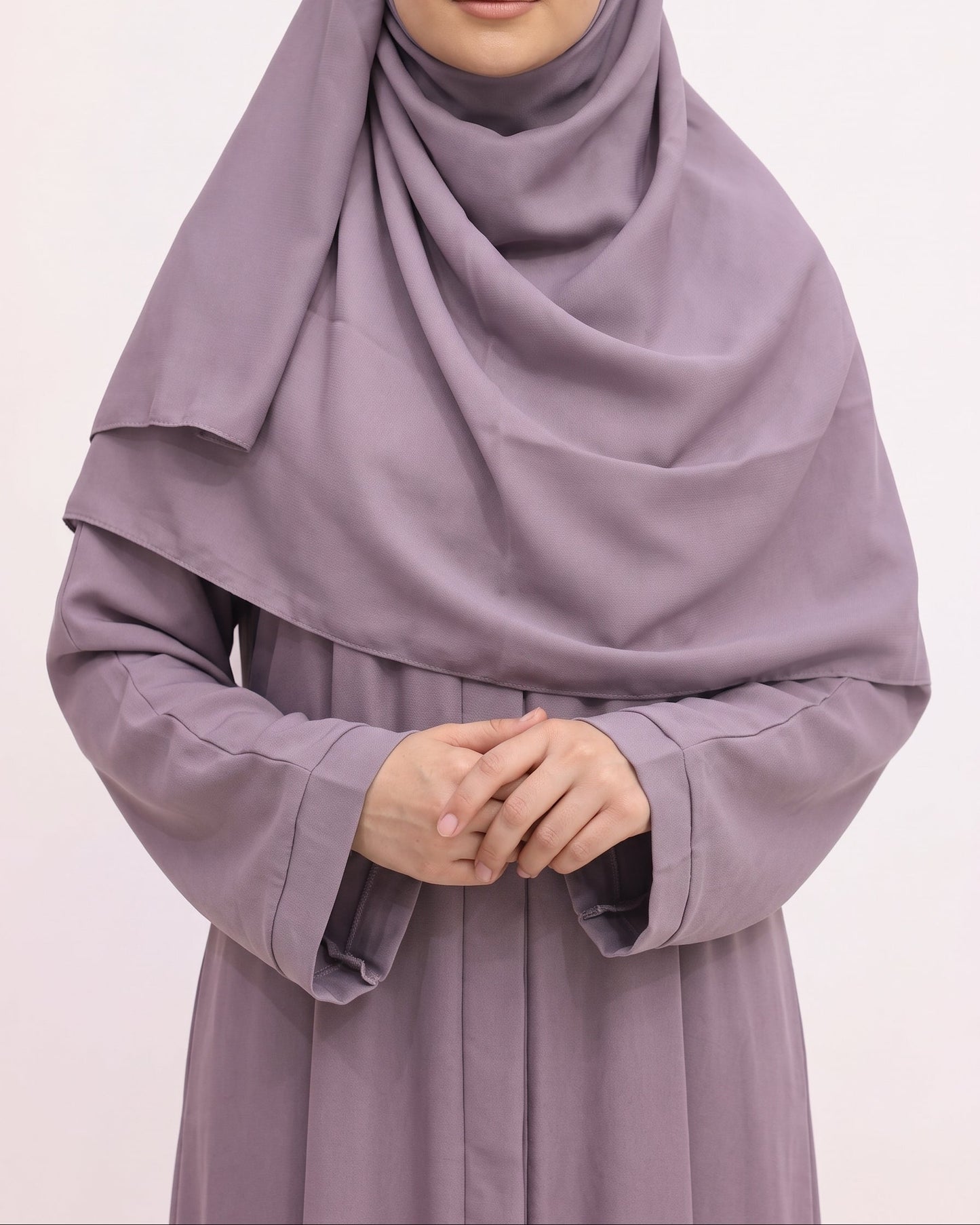 A woman wearing a draped dusty purple hijab and matching long-sleeve abaya against a plain light background.