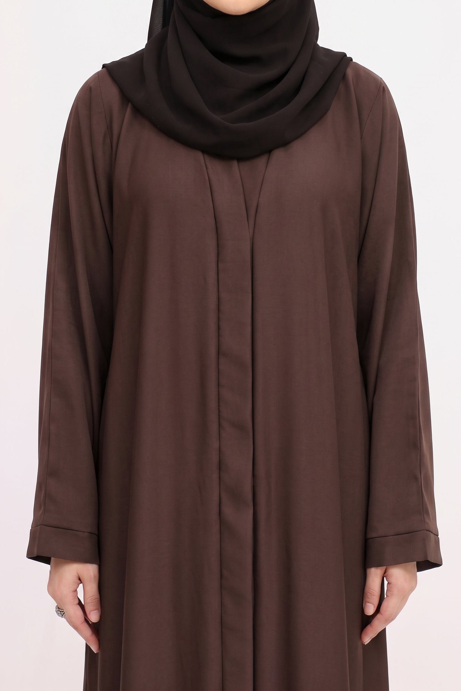 A woman wearing a long-sleeved brown abaya with a matching dark brown hijab, shown from the neck down against a plain white background.