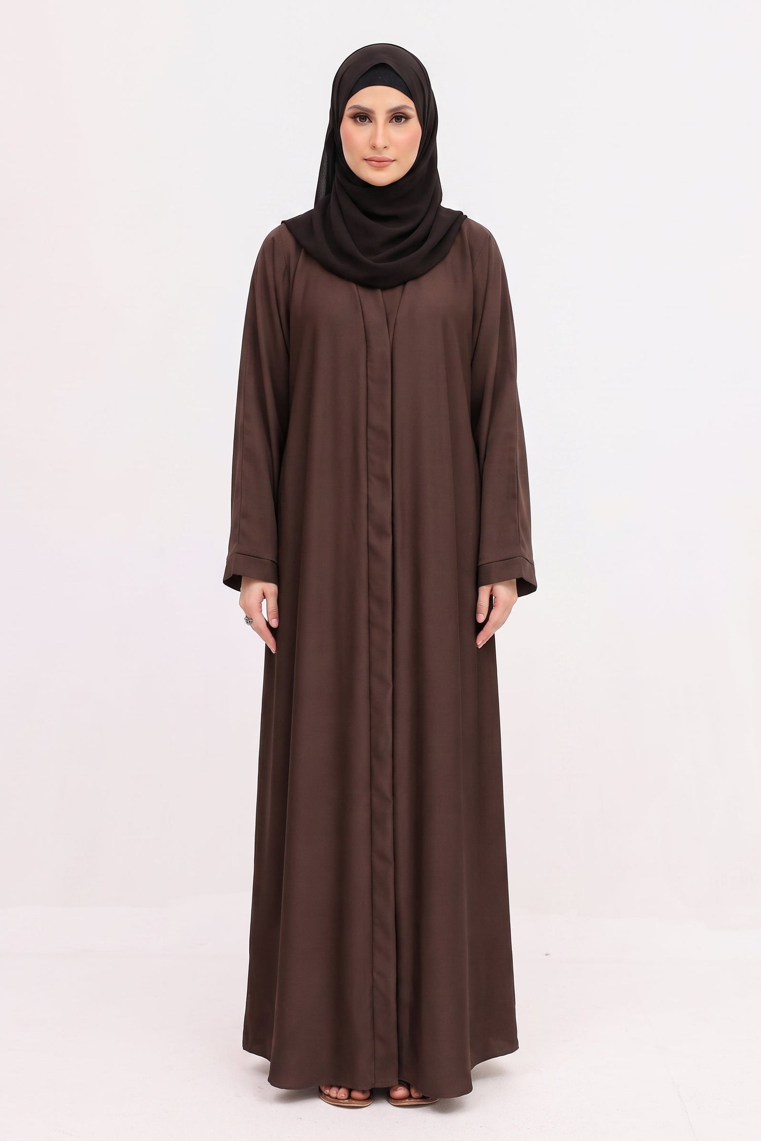 A woman modeling a long-sleeved brown abaya with a matching black hijab, standing against a plain white studio background.