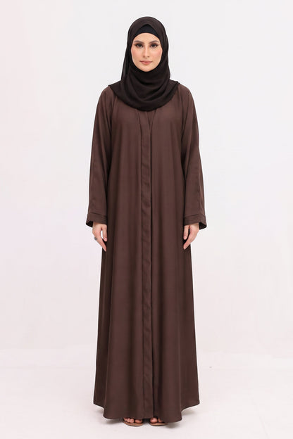 A woman modeling a long-sleeved brown abaya with a matching black hijab, standing against a plain white studio background.