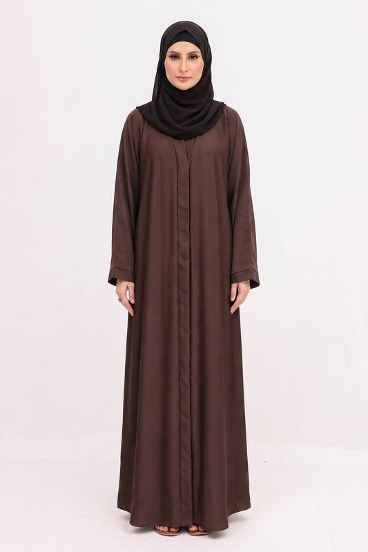 A woman modeling a long-sleeved brown abaya with a matching black hijab, standing against a plain white studio background.