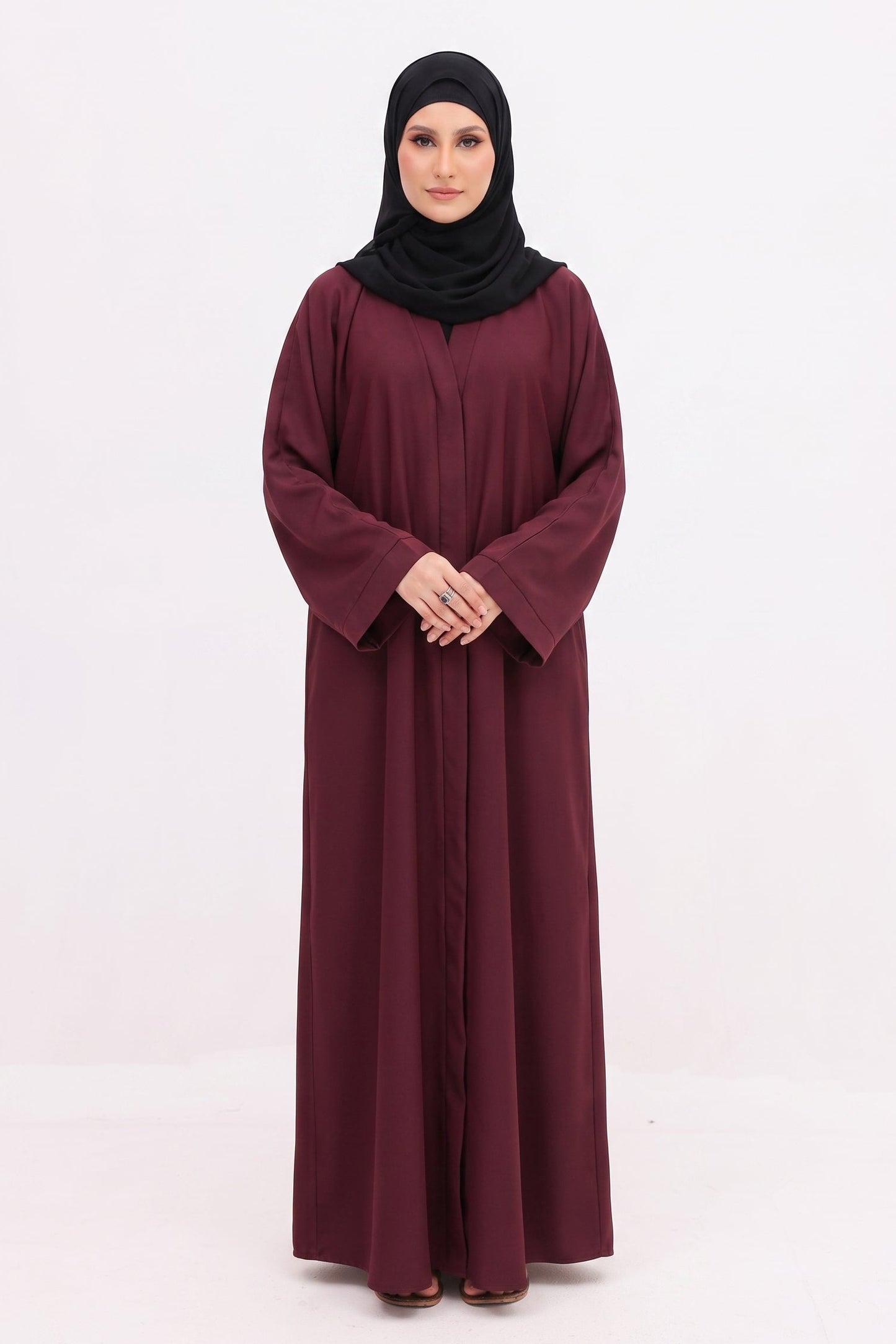 A woman wearing a long, loose-fitting maroon abaya with wide sleeves and a black hijab, standing against a plain white studio background.